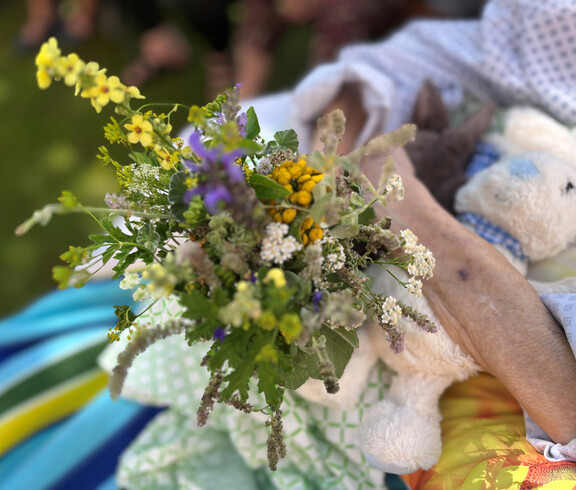 Ältere Frau hält liegend einen bunten Kräuterstrauß, daneben Stofftier auf der Decke
