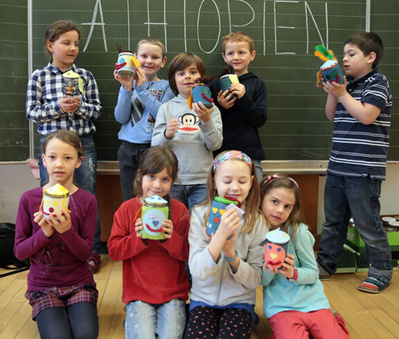 Junge Schulkinder sitzen und stehen in einer Klasse. Sie halten ihre Bastelfiguren in die Höhe und bestaunen sie.