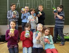 Junge Schulkinder sitzen und stehen in einer Klasse. Sie halten ihre Bastelfiguren in die Höhe und bestaunen sie.