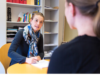 Zwei junge erwachsene Frauen sitzen an einem Tisch. Sie führen ein Gespräch und eine Frau notiert sich die Kernelemente der Besprechung. 