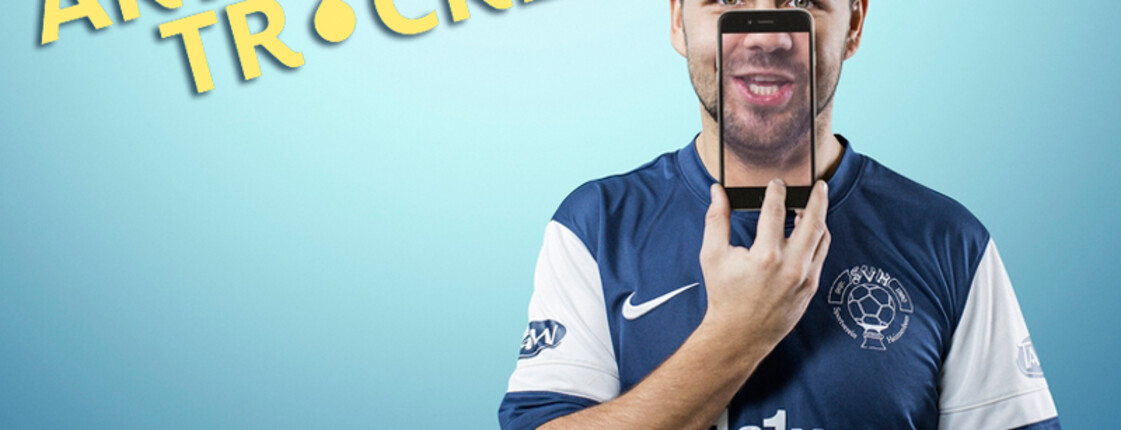 Ein junger Mann hält ein Handy über sein Gesicht, wobei man sein Gesicht durch das Handy sieht. Der Hintergrund ist hellblau und der Herr trägt ein blaues Fußballtrikot. 