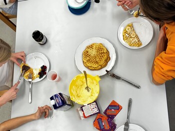 Ein weißer Tisch von oben, gebackene Waffeln und Toppings wie Bananen, Marmelade und Beeren, Teller mit belegten Waffeln und Hände der Mädchen von oben, die hier zusammen sitzen, Waffeln essen und sich unterhalten.