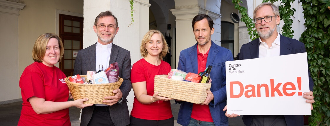 Mitarbeiter*innen der Caritas stehen in einem Innenhof und halten zwei mit Lebensmitteln gefüllte Körbe sowie ein Schild mit der Aufschrift "Danke!" in ihren Händen.