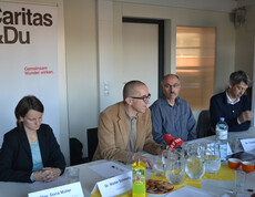 Eine Pressekonferenz mit Verantwortlichen der Caritas sitzen gemeinsam an einem langen Tisch. Ein ORF Mikrofon ist auf Walter Schmolly gerichtet. Im Hintergrund steht ein großes Plakat.