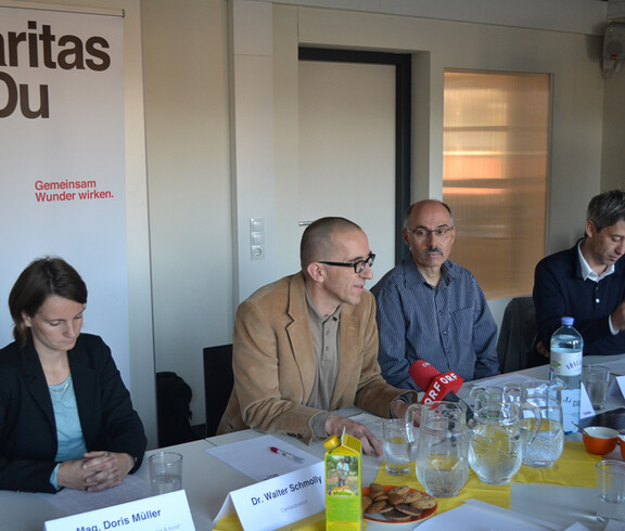 Eine Pressekonferenz mit Verantwortlichen der Caritas sitzen gemeinsam an einem langen Tisch. Ein ORF Mikrofon ist auf Walter Schmolly gerichtet. Im Hintergrund steht ein großes Plakat.