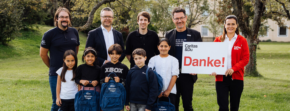 Acht Kinder mit Rucksäcken und sechs Erwachsene bei Caritas-Übergabeaktion im Grünen, Schild mit „Danke!“