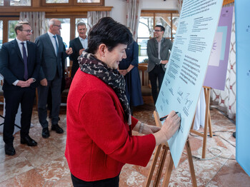 Eine Frau, die eine rote Jacke und einen Schal trägt, setzt ihre Unterschrift unter eine Charta, die auf einer Staffelei steht.