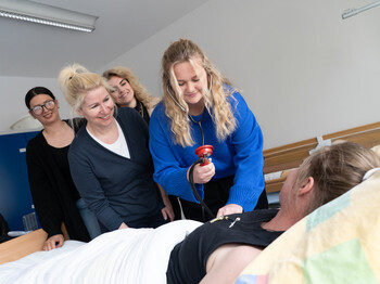 Eine Schülerin misst bei einer Mitschülerin den Blutdruck, die in einem Krankenbett liegt, die Lehrerin und Mitschüler*innen beobachten sie dabei.