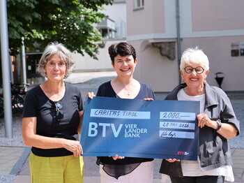 Drei Frauen stehen in einem Innenhof und präsentieren einen blauen Spendenscheck über 2.000 Euro.
