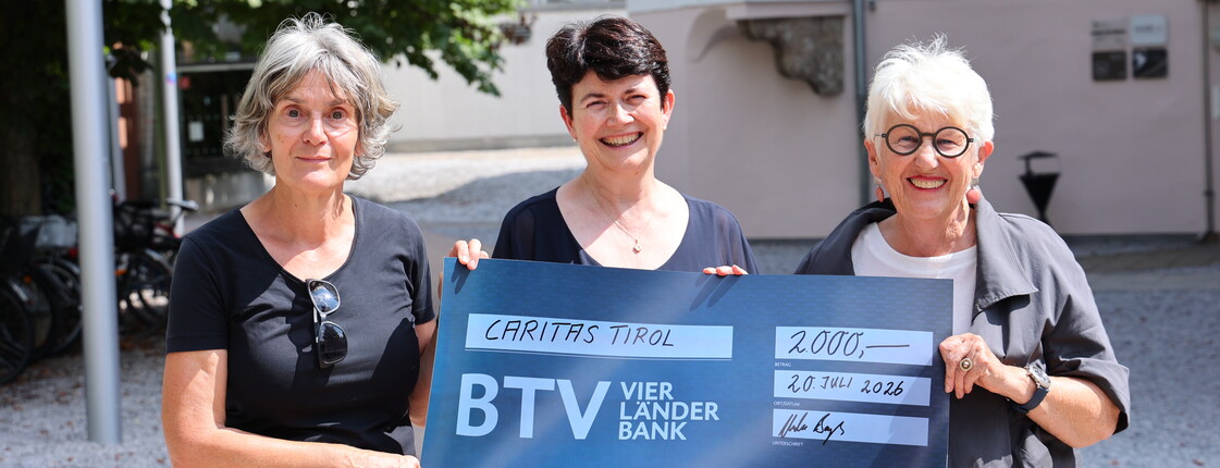 Drei Frauen stehen in einem Innenhof und präsentieren einen blauen Spendenscheck über 2.000 Euro.
