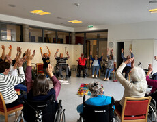 Senior*innen im Sitzkreis heben freudig beide Arme während einer Seelenyoga-Einheit.