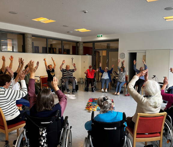 Senior*innen im Sitzkreis heben freudig beide Arme während einer Seelenyoga-Einheit.