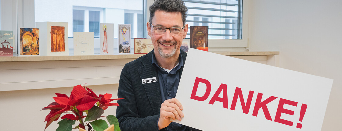 Ernst Sandriesser, Direktor der Caritas Kärnten, sitzt vor einem kleinen runden Tisch mit einem Weihnachtsstern und hält ein Schild mit "Danke" in der Hand.