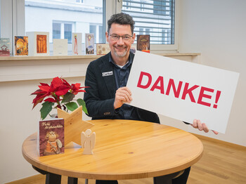 Ernst Sandriesser, Direktor der Caritas Kärnten, sitzt vor einem kleinen runden Tisch mit einem Weihnachtsstern und hält ein Schild mit "Danke" in der Hand.