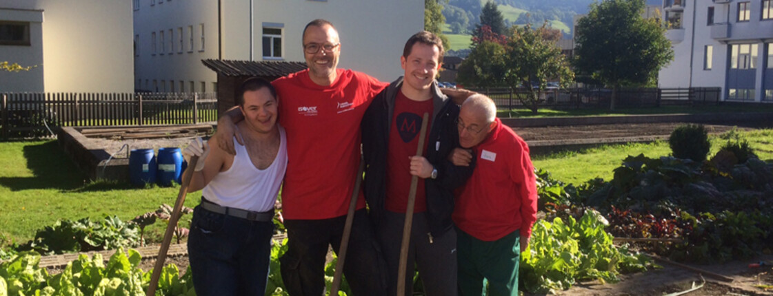 Mitarbeiter von Mondelez in Bludenz helfen im Garten der Werkstätte Bludenz bei schönem Wetter. Zwei Mitarbeiter und zwei Menschen mit Beeinträchtigung stehen im Garten zu einem Gruppenfoto zusammen. 