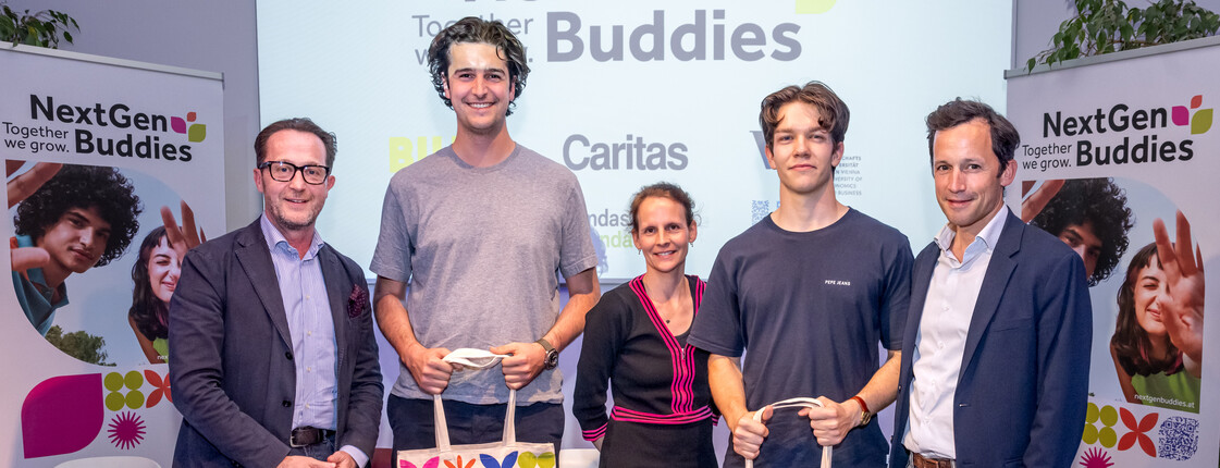Fünf Erwachsene stehen vor einer Leinwand auf die der Titel eines Projekts "Next Gen Buddies" projiziert wird. Zwei der Personen halten Tragetaschen mit dem Logo des Projekts in ihren Händen.