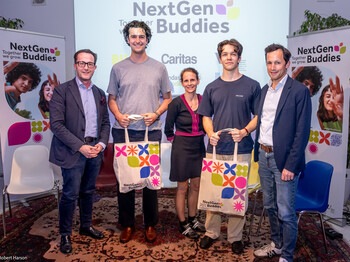 Fünf Erwachsene stehen vor einer Leinwand auf die der Titel eines Projekts "Next Gen Buddies" projiziert wird. Zwei der Personen halten Tragetaschen mit dem Logo des Projekts in ihren Händen.