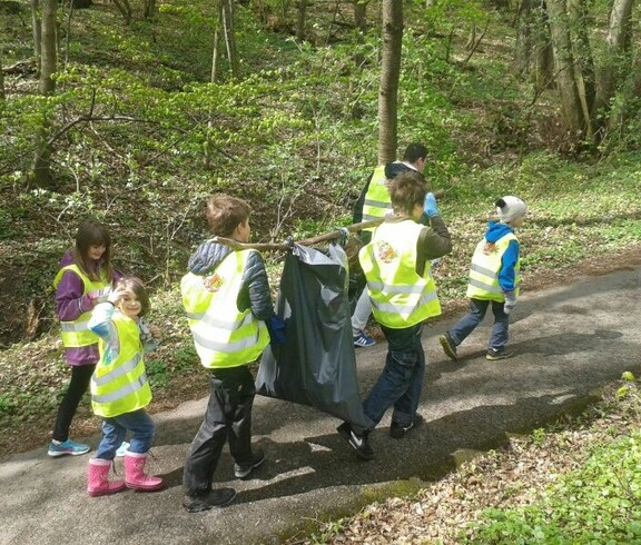 Kinder sammeln bei einem Waldputztag Abfälle entlang eines Waldwegs und tragen gemeinsam einen gefüllten Müllsack durch die Natur.