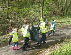 Kinder sammeln bei einem Waldputztag Abfälle entlang eines Waldwegs und tragen gemeinsam einen gefüllten Müllsack durch die Natur.