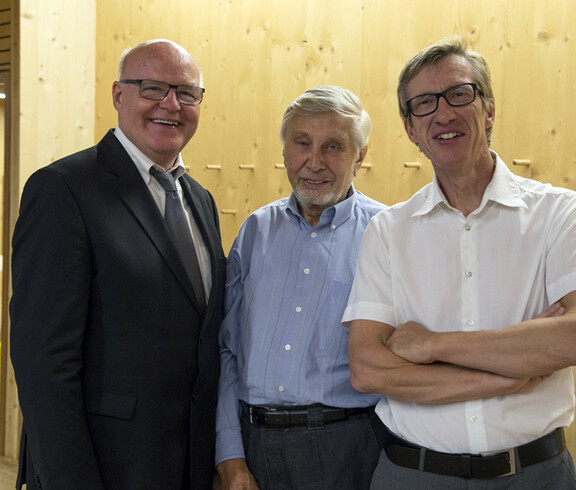 Drei Männer stehen im Vordergrund und lachen in die Kamera. Der ganz rechte Herr trägt ein weißes kurzes Hemd, der Herr in der Mitte ein blaues Hemd und der Herr links einen schwarzen Anzug. 
