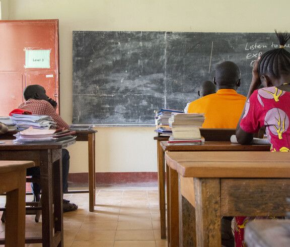 Drei Schüler sitzen von hinten an Holzschulbänken in einem einfachen Klassenzimmer im Südsudan, auf den Tischen liegen Schulbücher und Hefte.