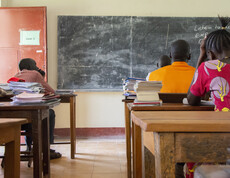 Drei Schüler sitzen von hinten an Holzschulbänken in einem einfachen Klassenzimmer im Südsudan, auf den Tischen liegen Schulbücher und Hefte.
