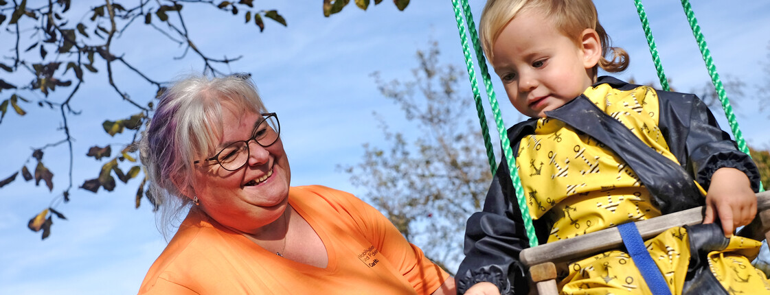 Eine kleines Mädchen sitzt auf dem Schoß einer Frau. Vor ihnen liegt ein Buch am Tisch, das sie sich gemeinsam ansehen.
