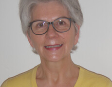 Das Foto zeigt eine Frau mittleren Alters. Sie trägt ein gelbes Oberteil, roten Lippenstift und eine schwarze Brille.