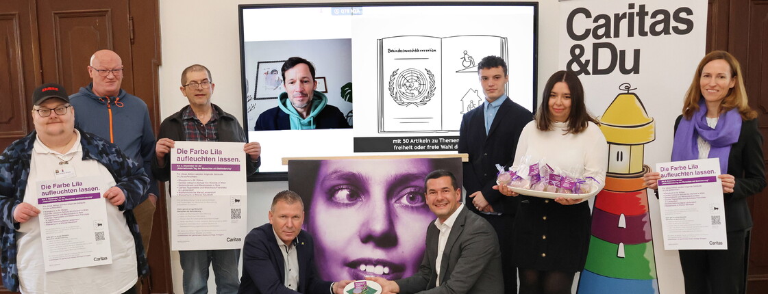Gruppenfoto von neun Personen bei einer Veranstaltung der Caritas. Zwei Männer knien vorn und halten gemeinsam ein lilafarbenes Herzsymbol. Weitere Personen stehen dahinter, drei von ihnen halten Plakate mit der Aufschrift „Die Farbe Lila aufleuchten lassen.“ Eine Frau trägt einen Tablett mit lilafarben verpackten Geschenken. Im Hintergrund ist ein Bildschirm mit einem Videocall und eine Illustration zur Menschenrechtskonvention zu sehen. Rechts steht ein Roll-up mit der Aufschrift „Caritas & Du“ und einem bunten Leuchtturm.