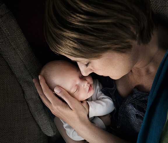 Eine junge Mutter hält ein neugeborenes in ihren Händen. Behutsam liegt der Kopf des Babys in einer ihren Händen. Beide haben die Augen geschlossen und es scheint, als genießen sie die Situation.