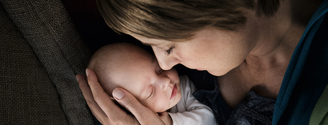 Eine junge Mutter hält ein neugeborenes in ihren Händen. Behutsam liegt der Kopf des Babys in einer ihren Händen. Beide haben die Augen geschlossen und es scheint, als genießen sie die Situation.
