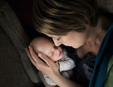 Eine junge Mutter hält ein neugeborenes in ihren Händen. Behutsam liegt der Kopf des Babys in einer ihren Händen. Beide haben die Augen geschlossen und es scheint, als genießen sie die Situation.
