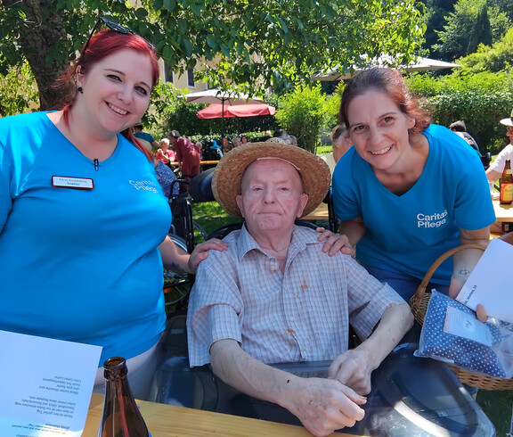 Bewohner mit Strohhut sitzt zwischen Mitarbeitenden beim Grillfest im Garten des St. Hemma Hauses.