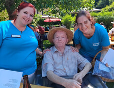 Bewohner mit Strohhut sitzt zwischen Mitarbeitenden beim Grillfest im Garten des St. Hemma Hauses.
