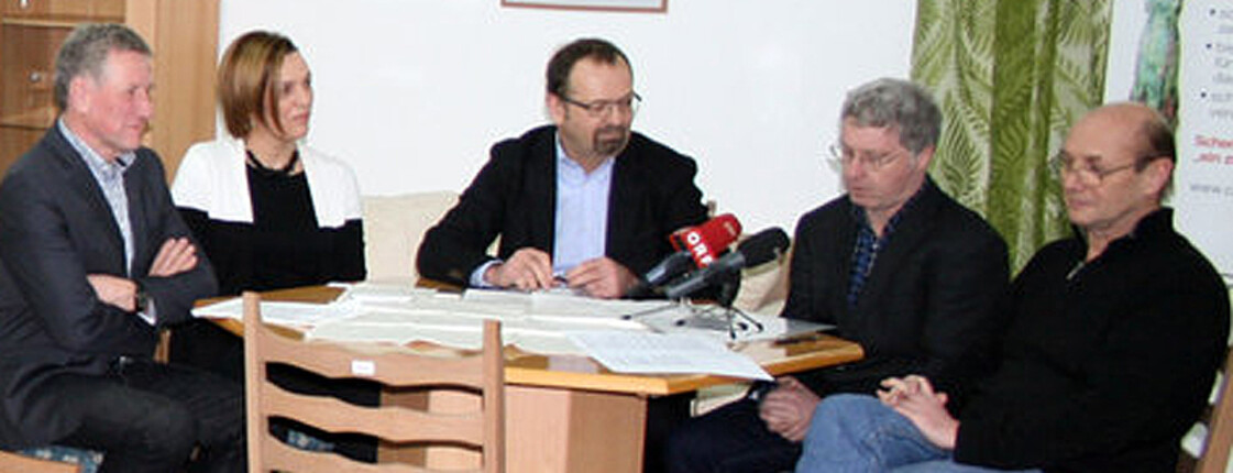 Fünf Menschen sitzen an einem runden Tisch beisammen. Auf dem Tisch stehen zwei Mikrofone.