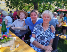 Bewohner*innen und Mitarbeitende sitzen gemeinsam an Festbänken im schattigen Garten des Pflegewohnhauses.