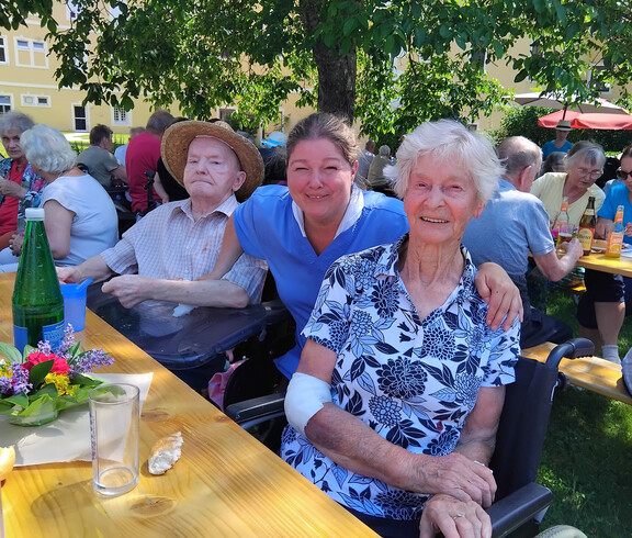 Bewohner*innen und Mitarbeitende sitzen gemeinsam an Festbänken im schattigen Garten des Pflegewohnhauses.