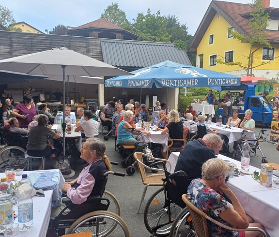 im Innenhof des Pflegewohnhauses sitzen zahlreiche Bewohnerinnen und Bewohner an festlich gedeckten Tischen, während im Hintergrund für Musik und Unterhaltung gesorgt wird.