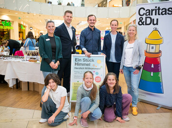 Mitarbeiter*innen der Caritas und drei kleine Mädchen stehen vor einem Marktstand in einem Einkaufzentrum und präsentieren mit Plakaten und Bannern die "Schule am Himmel".