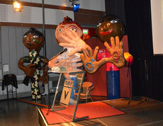 Ca. 2 Meter hohe Figuren stehen in einem größeren Saal. Sie repräsentieren Menschen unterschliedlicher Ehtnien. Im Vordergrund befindet sich eine Bühne. 