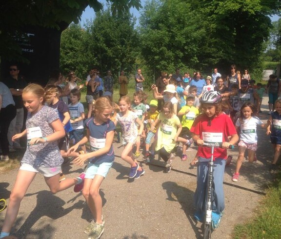 Kinder starten gemeinsam bei einer Sportveranstaltung im Freien. Teilnehmende laufen und fahren mit Rollern über die Strecke.