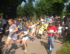 Kinder starten gemeinsam bei einer Sportveranstaltung im Freien. Teilnehmende laufen und fahren mit Rollern über die Strecke.