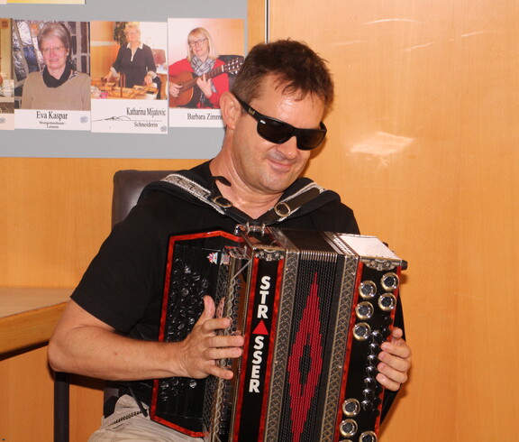 Ein blinder Mann sitzt auf einem Stuhl und spielt auf einer Ziehharmonika in den Farben Schwarz, Rot und Gold. Er trägt eine schwarze Sonnenbrille und wirkt konzentriert und zugleich entspannt beim Musizieren.