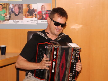 Ein blinder Mann sitzt auf einem Stuhl und spielt auf einer Ziehharmonika in den Farben Schwarz, Rot und Gold. Er trägt eine schwarze Sonnenbrille und wirkt konzentriert und zugleich entspannt beim Musizieren.