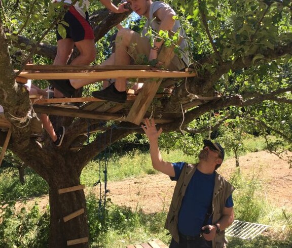 Mehrere Personen bauen gemeinsam eine Plattform in einem Baum und arbeiten unter Anleitung an einem handwerklichen Projekt im Freien.
