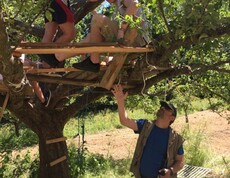 Mehrere Personen bauen gemeinsam eine Plattform in einem Baum und arbeiten unter Anleitung an einem handwerklichen Projekt im Freien.
