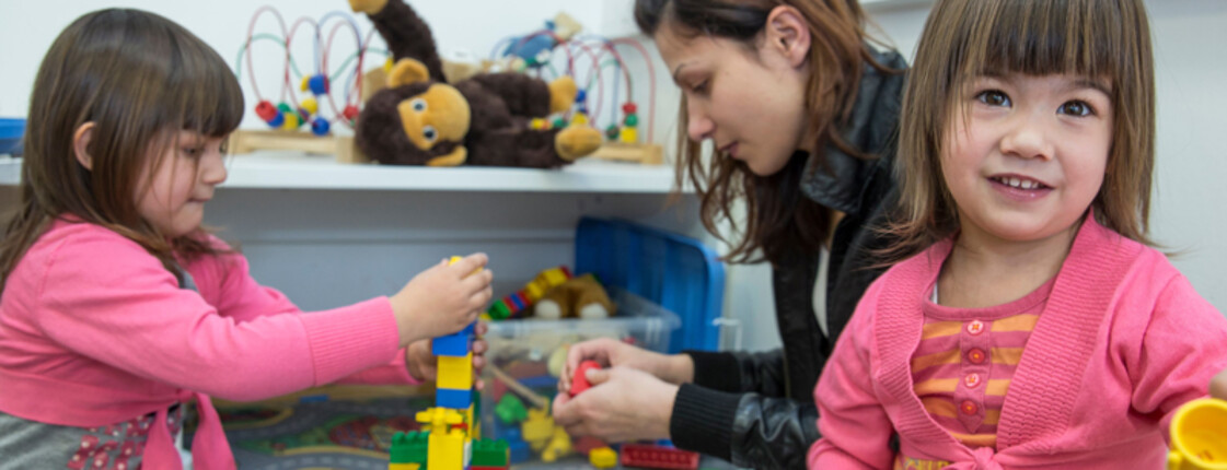 Eine junge Frau spielt mit zwei kleinen Mädchen am Boden Lego. Ein Mädchen im Vordergrund wirkt sehr fröhlich und lächelt. Im Hintergrund baut die junge Frau mit dem Mädchen einen Turm.