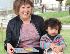 Eine ältere Dame sitzt mit einem kleinen Kind auf einer Bank. Die Frau strahlt und hält ein Buch in der Hand. Mit der anderen Hand hält sie das Kind im Arm.
