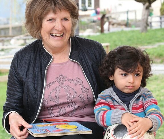 Eine ältere Dame sitzt mit einem kleinen Kind auf einer Bank. Die Frau strahlt und hält ein Buch in der Hand. Mit der anderen Hand hält sie das Kind im Arm.
