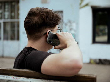 Ein junger Mann telefoniert mit dem Handy auf einer Bank vor einem weißen Haus.